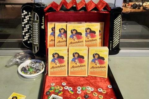 Accordion book sales display made from an accordion case and an old Hohner chromatic button accordion.
The left and right sides of the accordion are attached to the sides of the accordion case so that when you open it, the red velvet interior is where the bellows would be. Inside the Accordion case, where the Bellows of the accordion would be a neat display of my Accordion Revolution history of the accordion books. Below that in the open red-velvet-lined lid of the case, there are a selection of accordion themed buttons and stickers and pins.