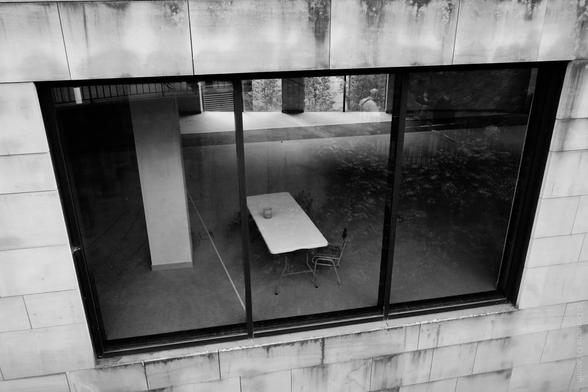 A black and white photo taken from outside a building in Edinburgh, Scotland, showing a large window with a view into a sparse room containing a single rectangular table, one chair, and a small object (possibly a candle or glass) on the table; the window glass reflects the exterior surroundings and faint figures, and the building facade is marked by weathering and stains.
