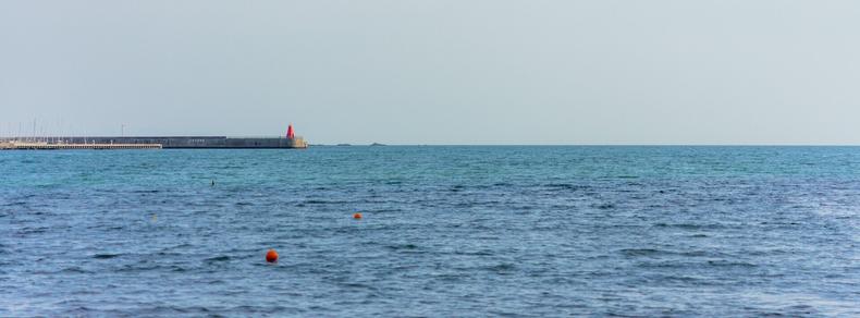 The image shows a calm sea with a few scattered orange buoys on the water. In the background, there's a breakwater with a red structure, possibly a lighthouse or marker, on top. The sky is clear and blends softly with the horizon.