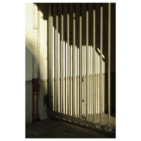 Photo einer mit einem Gitter-Tor verschlossenen Einfahrt. Tiefe Morgensonne taucht einen Teil des Gitters in warmes Licht und lässt das Tor senkrechte Schatten-Linien auf die dahinter liegende Wand werfen.
