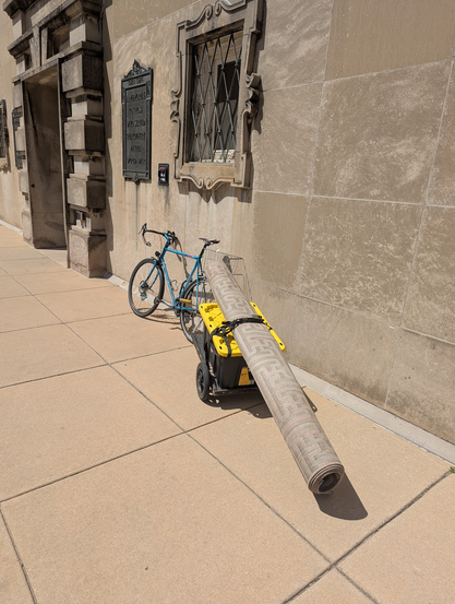 A rolled up rug strapped to the top of a crate on top a bike trailer. The end of the rug is hanging far over the end of the trailer and is dangerously close to the ground.