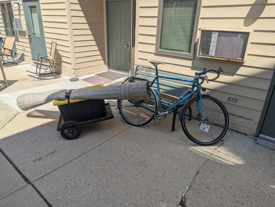 Side view of a rolled up rug strapped to the top of a crate on top a bike trailer. The rug sits diagonally on the trailer and is not quite as close to the ground.