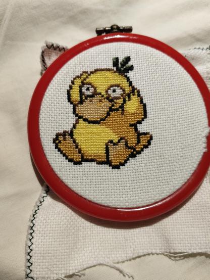 a cross stitch of the pokemon Psyduck, a yellow duck who is holding its head because it always has a headache.
It is on white weave in a red rubber frame.