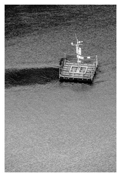 Schwarz-weiß Foto im Hochformat. Teleaufnahme einer Wasseroberfläche. Im oberen Drittel des Bildes ist eine kleine künstliche quadratische Insel. Auf allen vier Seiten ist ein Geländer, an einer eine Tür. In der Mitte ist eine Art Antenne. Das Wasser rundherum ist voller Miniaturen Wellen, die eine texturartige Oberfläche bilden.