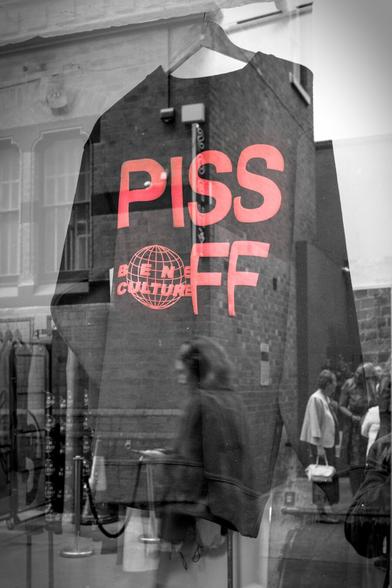 A black and white image of a jumper hanging in a shop window. The words on the jumper are the only colour in the picture - they are read and read "Piss *off"!

The shop window is reflecting the street scene. A woman is visible in the reflection, walking past, looking at here phone. Buildings and other people are also visible in the reflection.