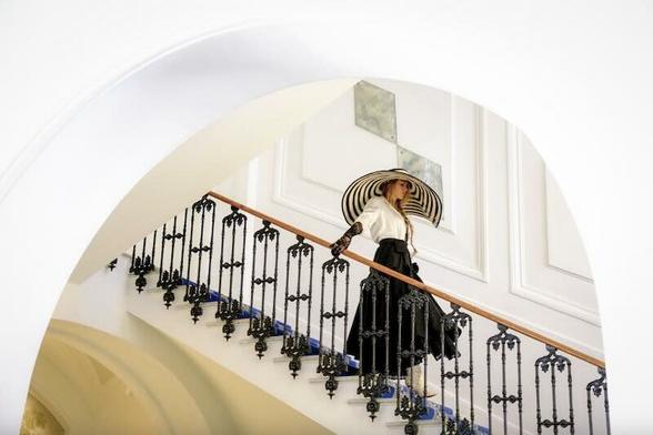 Blake Lively walks down a grand staircase in a huge black and white striped hat. She wears black gloves, a long black skirt, and a white blouse.