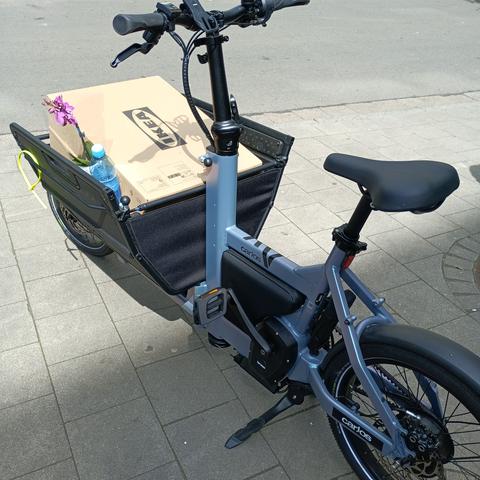 Ein hellviolettes Lastenrad mit Frontkorb, der mit Ikea Kartons gefüllt ist
