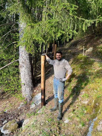 Am young men standing and smiliny near a wooden fencepost having on hand leaned on the top of the larch fence post. A large spruce tree in the background.
