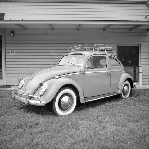 A black and white film photo. A classic Volkswagen Beetle is parked on some lawn in front of a building in Austin, Texas. Sunday, April 27, 2025.