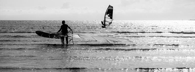 The image depicts a black and white scene of a beach with two individuals engaged in windsurfing. One person is close to the shore carrying a windsurfing board, while another is further out on the water riding the waves. The sea and sky create a serene backdrop.