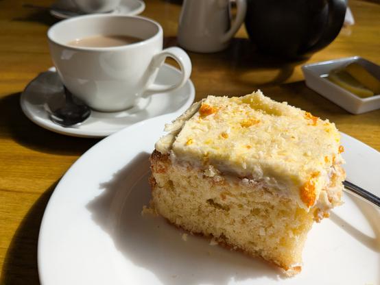 Colour photograph of a square chunk of sponge cake, topped with pale orange buttercream. Behind it is a cup of tea. All the crockery is white.