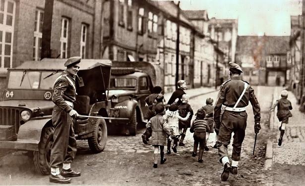 Schwarz-Weiß-Bild, das zwei Soldaten in Militäruniformen zeigt, die eine Gruppe von Kindern beaufsichtigen, die durch eine gepflasterte Straße laufen. Ein Militärjeep ist in der Nähe geparkt, und historische Gebäude säumen den Hintergrund. Die Szene vermittelt ein Gefühl von Nostalgie und vielleicht einen Moment des kontrollierten Spiels