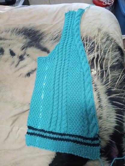 A shaped panel of knitted fabric with cables in sky blue with dark blue stripes at the bottom.