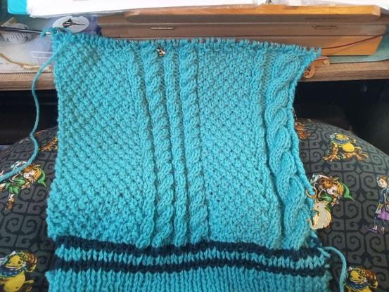 A panel of knitted fabric with cables in process. It's sky blue with dark blue stripes at the bottom.
