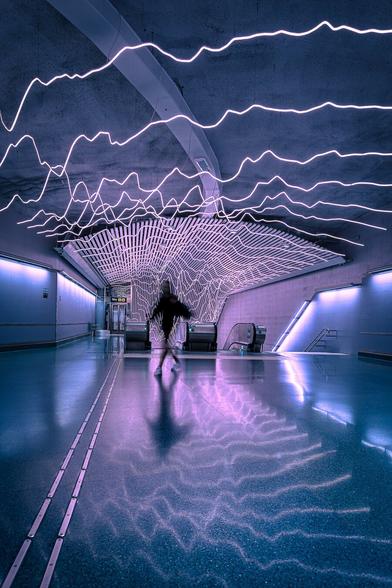 A girl is crossing a dark hall in blueish tones under zigzagging pink neon lights after coming up an escalator at Odenplan metro station in Stockholm. Her tiny dark silhouette is slightly blurry due to a long exposure and his reflected on the shiny floor.