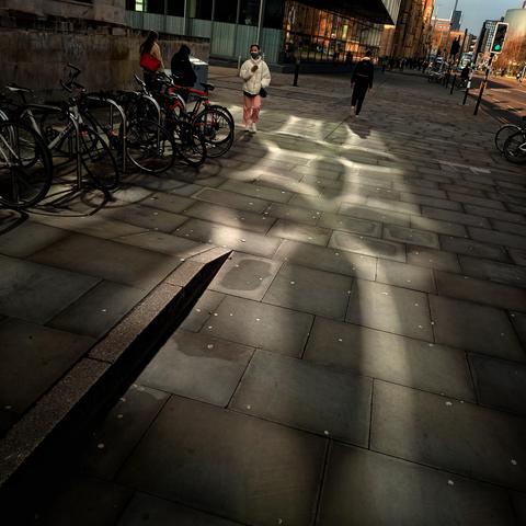 reflection of morning light on a building making nice patterns on the ground, street with people and bikes.
