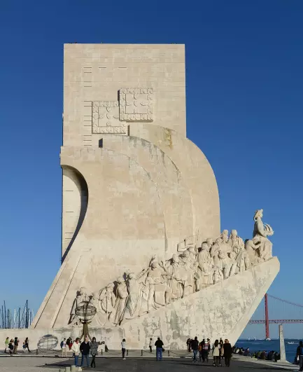 The image depicts the Monument to the Discoveries, located in Lisbon, Portugal. This monument honors the Portuguese explorers of the Age of Discoveries. It features a stylized ship with various figures representing explorers, navigators, and other notable individuals from that era. The structure is made of stone and stands against a clear blue sky, with people visible in front of it, likely admiring the monument. In the background, you can see part of the waterfront and boats.

Image Credits: Wikimedia / Alvesgaspar / CC BY-SA 4.0