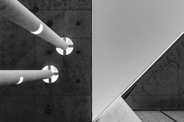 Abstract black and white photo of modern architecture showing concrete columns extending through circular ceiling openings on the left, and an angular corner of a building against a pale sky on the right.