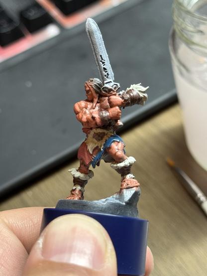 The HeroQuest Barbarian on a paint handle. The body is base colored with the hair and sword missing so far. It is shown from its left side.