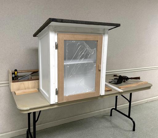 Little Free Library box sitting on a table with a toolbox to the back and a hand drill to the side. The box is approximately 28" tall and 24" wide made with plywood and thin pieces of pine trim at the corners. The roof slants from front to back and is covered with asphalt roofing material. The roof overhangs about 4" in front and 2" on the sides and back. The door is made with white oak boards supporting plexiglas. The oak will have a clear finish. Through the plexiglas a shelf is visible. The shelf is mounted 13" from the bottom - enough room for most picture books.