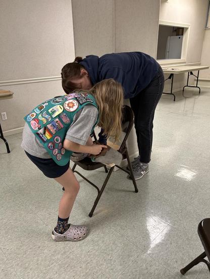 A woman and a girl leaning over a chair. The girl is wearing a green vest with many Girl Scout badges and is sawing a piece of the trim.