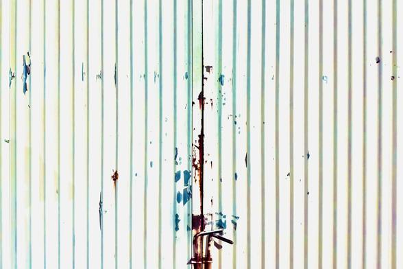 A large, white, metallic gate stands closed, with thin vertical shadows running down it like stripes, along with rust and teal paint chipping away at its outer coat.