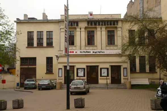 The image depicts a building that appears to be a theater, indicated by signs and the architecture of the structure. It features a façade with large windows and an entrance that has posters or signage displayed. There are cars parked in front, and it looks like there are some decorative planters or benches in the area. The overall setting seems to be a public space, likely in an urban environment.

Image Credits: Wikimedia / Gapcior / Public domain