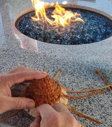 Close-up of hands weaving a cedar bark mini hat on a gray speckled stone countertop. Long, thin strips of brown material project from the unfinished basket. In the background, a modern fire pit filled with blue glass has flames burning. Pins with red and green heads are visible near the weaving.