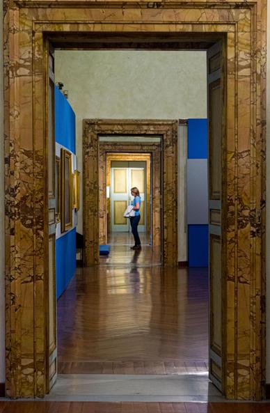 View through several open doors in a row in a museum. The edges of the door frames are very wide and made of polished, flamed brown wood. The door frames of the first door surround the entire photo like a picture frame. Behind it in the first room you see that the left wall is painted bright blue with a horizontal wide white strip in the middle on which paintings with wooden frames are hung. You see a total of four identical doors, which become smaller and smaller due to the perspective. With a woman in the back wearing a blue T-shirt who is looking at the paintings on the left.