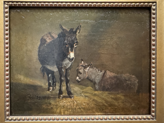 photo of the painting in gold frame on display at museum: small oil portrait of two donkeys in what appears to be a stall (brown wall, hay floor), one to left standing face forward and one to left lying down side profile facing left  “Constable spent the winter of 1815–16 in East Bergholt, Suffolk, England, with his dying father. Making the most of the unusually warm weather and his proximity to rural subjects, he painted these donkeys on February 29, 1816. Worked on a small scrap of canvas, the sketch served as a preparatory study that Constable used when adding donkeys to three landscapes: a wood scene in autumn, a view of Hampstead Heath, and a path outside the village of Stoke-by-Nayland. At an unknown date, probably following the artist’s death, the sketch was mounted on a piece of wood and the paint extended about an inch in all directions.”