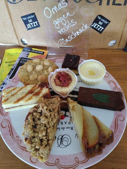 Auf einer runden Kuchenplatte mit rosafarbenem Rand thronen verschiedene Leckereien (im Uhrzeigersinn): Millionaire's Shortbread, Lemon Curd Cheesecake, Brownie mit frischer Minze aus meinem Garten, gestürzter Birnenkuchen mit Tonkabohne, 1/2 Apfel-Karamell-Törtchen mit Haferflocken-Zimt-Streuseln, Caramel Shortbread, Keks mit weißer Schoki & Macadamianüssen und in der Mitte ein Cheesecake mit Rhabarberkompott. 
Dahinter steht ein Schild, auf dem mit weißer Schrift auf den Klönschnack der Omas gegen rechts hingewiesen wird. 
Links davon liegen verschiedene Flyer mit Informationen zu Veranstaltungen in der Umgebung.