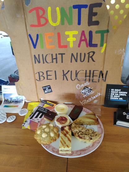 Auf einer runden Kuchenplatte mit rosafarbenem Rand thronen verschiedene Leckereien (die genaue Beschreibung findet sich unter dem ersten Foto).
Dahinter steht ein Pappschild (mein Schild für Demonstrationen), auf dem in großen Lettern Folgendes steht: Bunte Vielfalt, nicht nur bei Kuchen. 
Links und rechts vom Schild liegt verschiedenes Informationsmaterial von den Omas gegen rechts und der Aktion *AfDnee*.