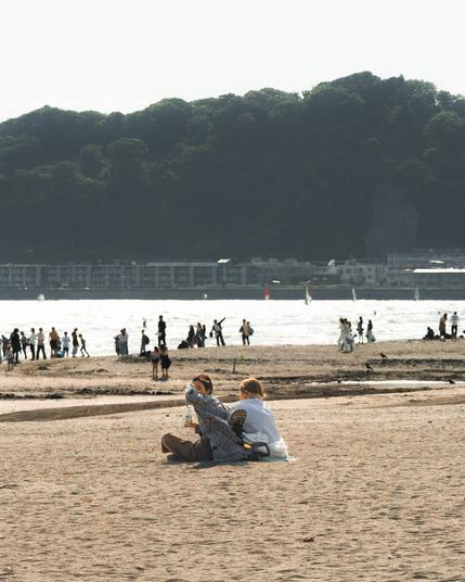 The image shows a beach scene with people relaxing and walking on the sand. In the foreground, a couple is sitting on the beach with some personal items next to them. In the background, more people can be seen near the shoreline. There are hills or a forested area across the water, and some buildings are visible along the coast. The atmosphere appears calm and sunny.