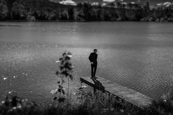 Monochrome shot of person stood on wooden jetty at pond edge watching the water. Trees line the far shore