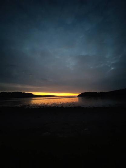 Sunset has created a small slit of orange glow on the horizon. It is pinned down by a large area of fluffy grey clouds. The water carries the glow toward the camera before it reaches the dark shore.