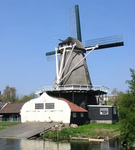 The image features a traditional windmill set against a clear blue sky. The windmill has a large, cylindrical body with a thatched roof and prominent blades. In front of the windmill is a structure resembling a barn or storage building, which is likely used for equipment or maintenance related to the windmill. The scene includes a body of water reflecting the windmill and its surroundings, adding to the picturesque rural landscape.

Image Credits: Wikimedia / Original uploader was Hans de Kroon at nl.wikipedia / CC BY-SA 3.0