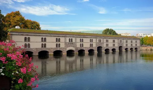 The image shows a building reflecting in a body of water, surrounded by greenery and flowers. The architecture appears to be historic, with arched windows and a green roof. The setting looks peaceful, possibly in an urban area with blue skies and some clouds.

Image Credits: Wikimedia / Claude TRUONG-NGOC / CC BY-SA 3.0