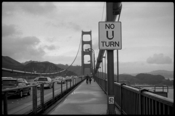 Black and white photography from the sidewalk of a bridge in front of a "No U turn" sign. The sky is cloudy.