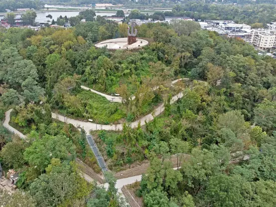The image appears to show a green, hilly area with winding paths and a circular structure at the top. The landscape is heavily wooded, suggesting a park or nature reserve. There seems to be some urban development in the background, indicating proximity to a city. The circular structure features a large sculpture or monument. The overall atmosphere seems tranquil and natural.

Image Credits: Wikimedia / Emptywords / CC BY-SA 4.0