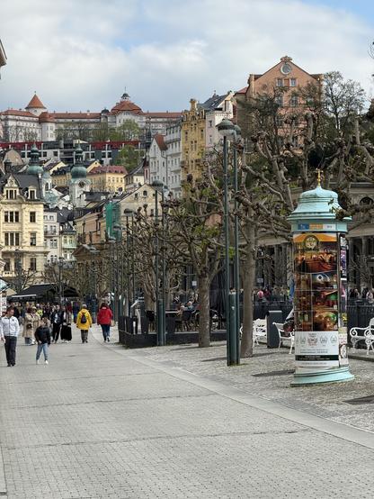 Das Bild zeigt eine Straße in einer tschechischen europäischen Stadt, gesäumt von Bäumen mit kahlen Ästen und historischen Gebäuden. Im Vordergrund verläuft eine gepflasterte Straße, auf der einige Fußgänger zu sehen sind.  Im mittleren Bereich steht ein Werbesäule mit mehreren Anzeigen, darunter Werbung für ein Spa und Bier.  Die Architektur der Gebäude im Hintergrund ist vielfältig und deutet auf einen alten Stadtkern hin. Der Himmel ist leicht bewölkt. Das Gesamtbild wirkt ruhig und strahlt eine gewisse historische Atmosphäre aus.