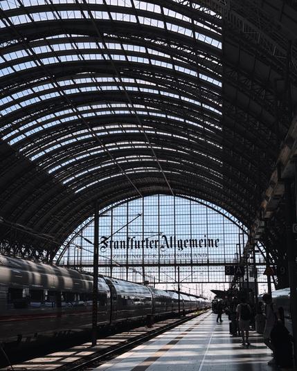 A train station with a high, arched glass roof. A long train is visible on the platform, and people are waiting or walking
