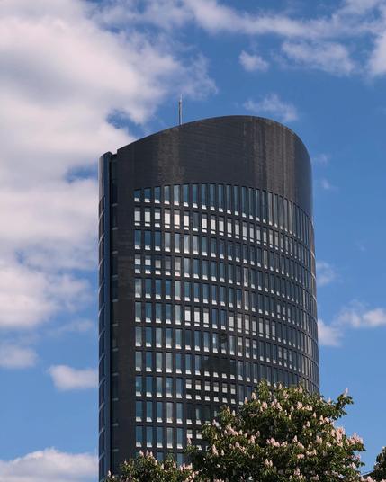 A modern high-rise building with a curved top, featuring a facade of dark glass windows. Part of a tree with pink flowers is visible in the foreground, set against a blue sky with scattered clouds