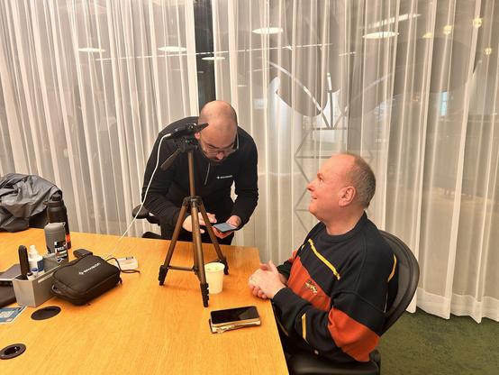 A man in an Envision-branded black sweater sets up a smartphone on a tripod, while another man in a striped sweater sits and smiles.
