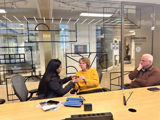 A woman demonstrates a device to a woman in a yellow sweater, while another man watches. They are seated in a glass-walled conference room labeled “Rectangle Conference Room.”