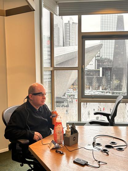 A man wearing smart glasses sits by the window with a view of a cityscape, holding a Fanta can. Various gadgets and snacks are on the table in front of him.