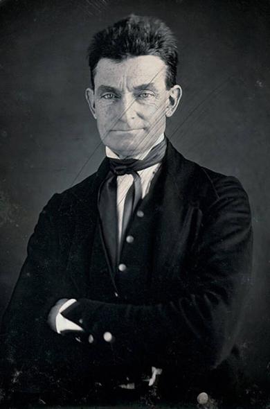 Black and white photo of Abolitionist John Brown wearing a suit and scowling with arms crossed against a gray background