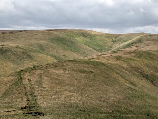 Colour photograph looking down a long narrow valley with hills rising steeply on both sides and at the end. It is covered in grass. A footpath is visible along the right hand hilltop twisting away into the distance. The sky is cloudy.