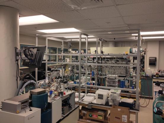 My little piece of the USian scientific enterprise. A small laboratory about 7 meters by 5 meters, packed with equipment. In the right front, you see green compressed gas bottles, across from that in the left bottom there is an isotope ratio mass spectrometer, and there is a homemade vacuum line and pyrolysis system set up across the background. You cannot see the fume hood and analytical balances tucked in behind the vacuum separations line. Fluorescent light fixtures line the ceiling.