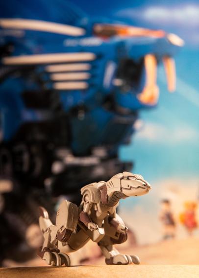 A small, mechanical dinosaur figure is in the foreground, with a blurred blue robotic creature looming behind it. The background features a colorful, sunny scene with indistinct figures, suggesting a playful environment.