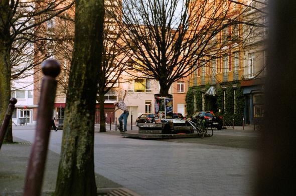 Colored photography of a skater performing a backsidd 5-0 grind on a small marble platform in a city place.